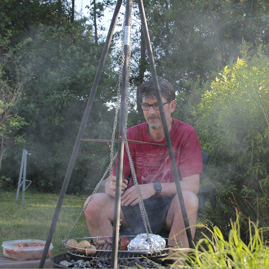 Ein Mann sitzt im Freien neben einem Grill, der über einem Feuer hängt. Er trägt ein rotes T-Shirt und eine Brille. Auf dem Grill liegen verschiedene Speisen. Im Hintergrund sind grüne Pflanzen und eine ruhige Gartenumgebung sichtbar. Rauch steigt vom Grill auf.