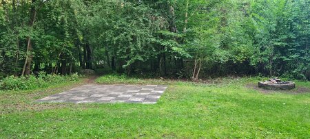 Eine grüne Wiese im Wald mit einem großen Schachbrett aus Steinplatten in der Mitte und einer Feuerstelle aus Steinen rechts im Bild. Im Hintergrund sind Bäume und Büsche zu sehen.