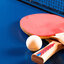 Tischtennisschläger mit roter Schlagfläche und Tischtennisball liegen auf einem blauen Tisch. Im Hintergrund ist ein Netz sichtbar.