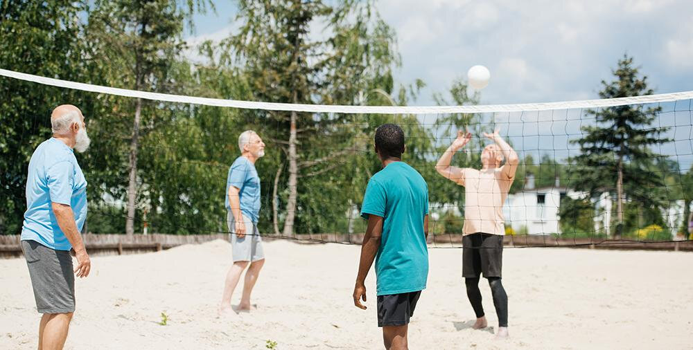 Vier Männer spielen Volleyball auf einem Sandplatz. Sie sind in sportlichen T-Shirts gekleidet und konzentrieren sich auf den Ball, der über das Netz fliegt. Im Hintergrund sind Bäume und Gebäude sichtbar.