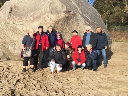 Eine Gruppe von zwölf Personen steht vor einem großen Felsen auf sandigem Boden. Einige tragen rot und schwarz, während andere helle Kleidung gewählt haben. Die Sonne scheint, und der Hintergrund zeigt Bäume und Sträucher. Die Stimmung wirkt freundlich und entspannt.