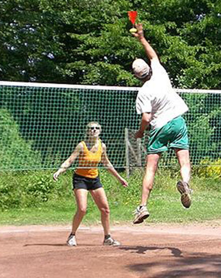 Ein Mann springt, um einen roten Becher über einem Netz zu erreichen, während eine Frau in kurzer Sportkleidung zuschaut. Der Hintergrund zeigt Bäume und eine Sandplatzfläche.