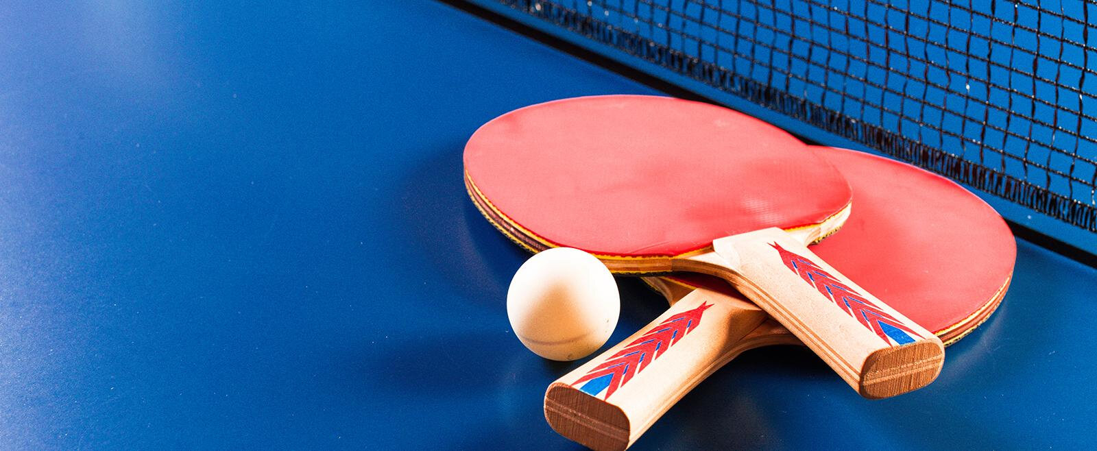 Zwei Tischtennis-Schläger mit roten Belägen liegen auf einer blauen Spielfläche, daneben ist ein weißer Tischtennisball. Im Hintergrund ist ein Netz sichtbar.