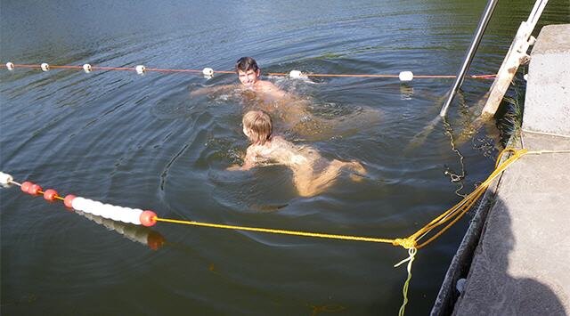 Zwei Kinder schwimmen in einem Gewässer, umgeben von einem Schwimmseil. Der vordere Junge ist blond und taucht, während der andere hinter ihm schwimmt und lacht. Am Rand des Wassers sind Steine und ein Metallgeländer sichtbar.