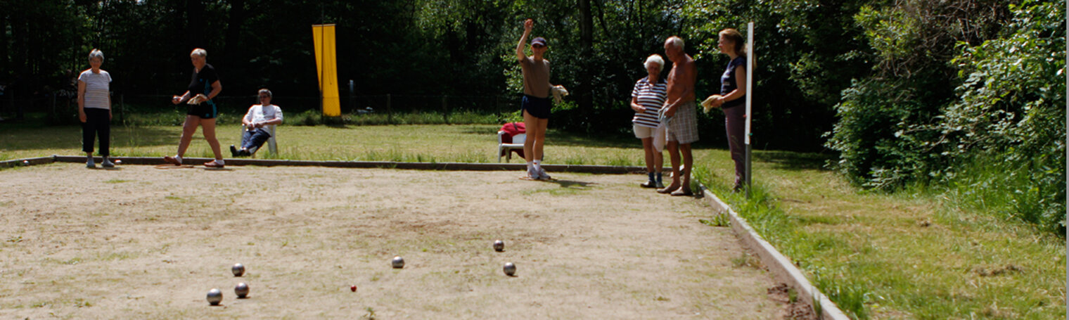 Auf einem Bouleplatz spielen mehrere Personen. Eine Spielerin wirft eine Kugel, während andere zuschauen. Im Hintergrund sind Bäume und eine Wiese zu sehen. Einige Personen sitzen und stehen in der Nähe der Spielfläche.