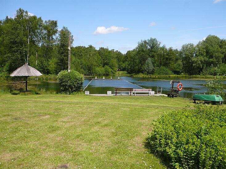 Ein ruhiger See mit umgebenden Bäumen und Grünflächen. Im Vordergrund steht eine Bank und ein kleiner Pavillon, während im Wasser Bojen und ein Rettungsring sichtbar sind. Ein Boot liegt am Ufer. Der Himmel ist blau mit einigen Wolken.