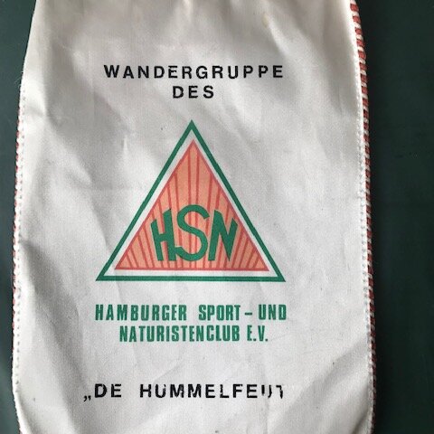 Eine weiße Tragetasche mit rot-weißer Schnur. Auf der Tasche steht "WANDERGRUPPE DES HSN HAMBURGER SPORT- UND NATURISTENCLUB E.V." in schwarzer Schrift. In der Mitte befindet sich ein grünes Dreieck mit dem roten Text "HSN". Unten steht "DE HUMMELFEUIT".