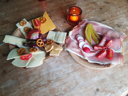 Auf einem Holzbrett präsentiert sich eine Käse- und Wurstplatte. Links sind verschiedene Käsestücke mit Trauben, Tomaten und Brezeln. Rechts liegt Schinken mit Radieschen und Essiggurken. Eine Kerze sorgt für gemütliche Beleuchtung im Hintergrund.