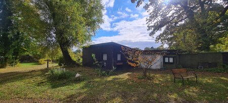 Ein schwarzes Haus steht in einer von Bäumen umgebenen, grünen Landschaft. Einige Blätter liegen auf dem Boden. Ein kleiner Holzstuhl befindet sich im Vordergrund. Der Himmel ist teilweise bewölkt und die Sonne scheint durch die Äste der Bäume.