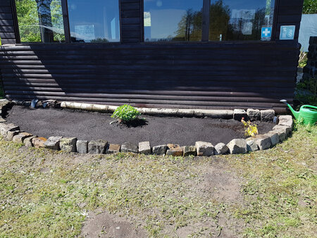 Eine von Steinen umschlossene Gartenfläche mit frischem Boden. Darin ist eine kleine Pflanze sichtbar, umgeben von Erde. Im Hintergrund steht ein dunkles Holzgebäude mit Fenstern, und rechts ist eine grüne Gießkanne zu sehen.