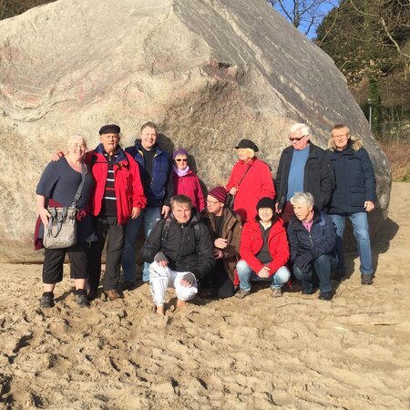 Eine Gruppe von etwa 15 Personen steht vor einem großen Felsen auf sandigem Boden. Die Mitglieder der Gruppe tragen unterschiedliche Kleidungsstücke, überwiegend in Rot und anderen Farben. Es ist sonnig, und einige Personen lächeln in die Kamera.