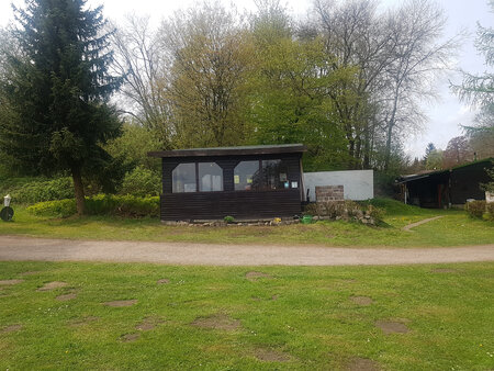 Eine einfache Holzstruktur mit großflächigen Fenstern steht auf einem grünen Rasen. Im Hintergrund sind Bäume und weitere Gebäude zu sehen. Der Weg vor der Struktur ist unbefestigt und von Gras umgeben. Es ist bewölkt und die Umgebung wirkt ruhig und naturnah.
