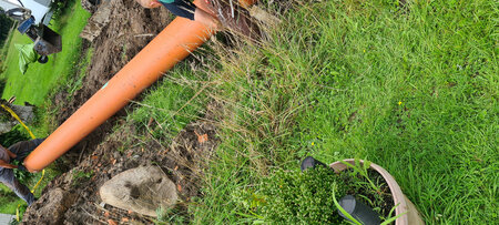 Ein Arbeiter gräbt in einem Garten, während er an einem orangefarbenen Rohr arbeitet. Im Vordergrund steht ein Blumentopf mit einer Pflanze. Der Boden ist erdig und umgeben von grünem Gras und vereinzelten Steinen.