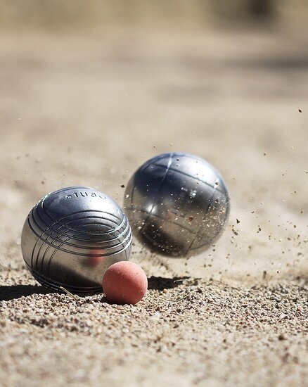Zwei metallene Kugeln rollen auf einem Sandplatz in Richtung einer kleineren roten Kugel. Der Sand wirbelt auf, während die Kugeln sich bewegen.